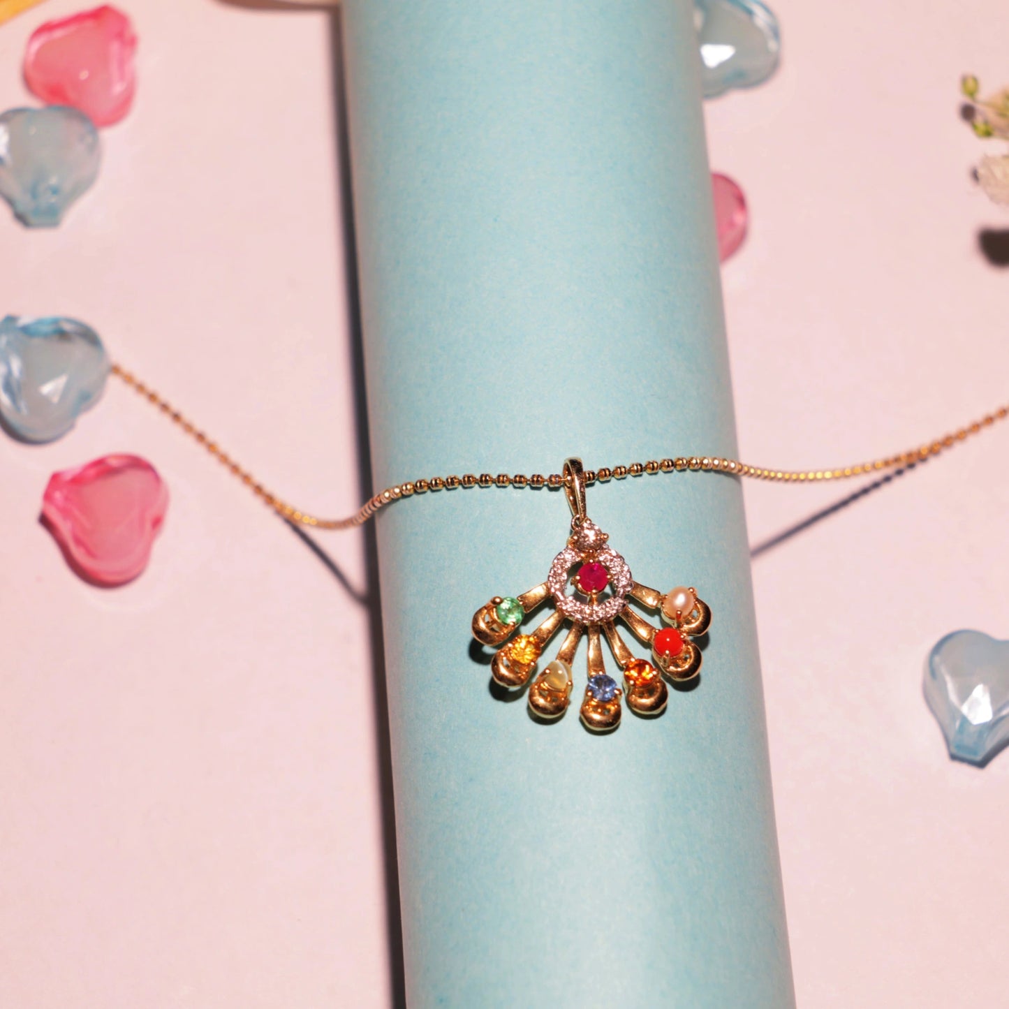 Artistic lifestyle shot of a vibrant Navratna pendant by jnjbymadhuri, displayed on a delicate gold chain over a pastel blue cylinder, surrounded by soft-focus rose petals and a dreamy pink backdrop—highlighting the pendant’s nine colorful gemstones and elegant fan-shaped gold frame.