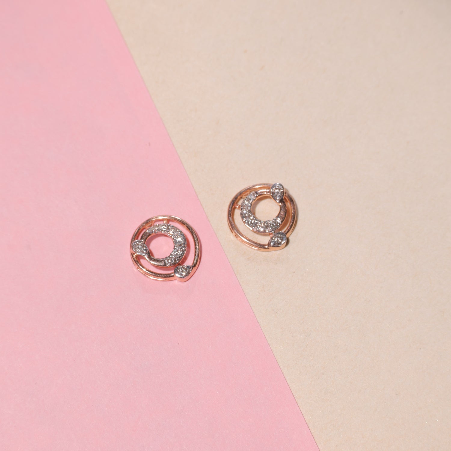 "Rose gold circular earrings with diamond accents arranged on a diagonally split pink and beige background, from jnjbymadhuri."