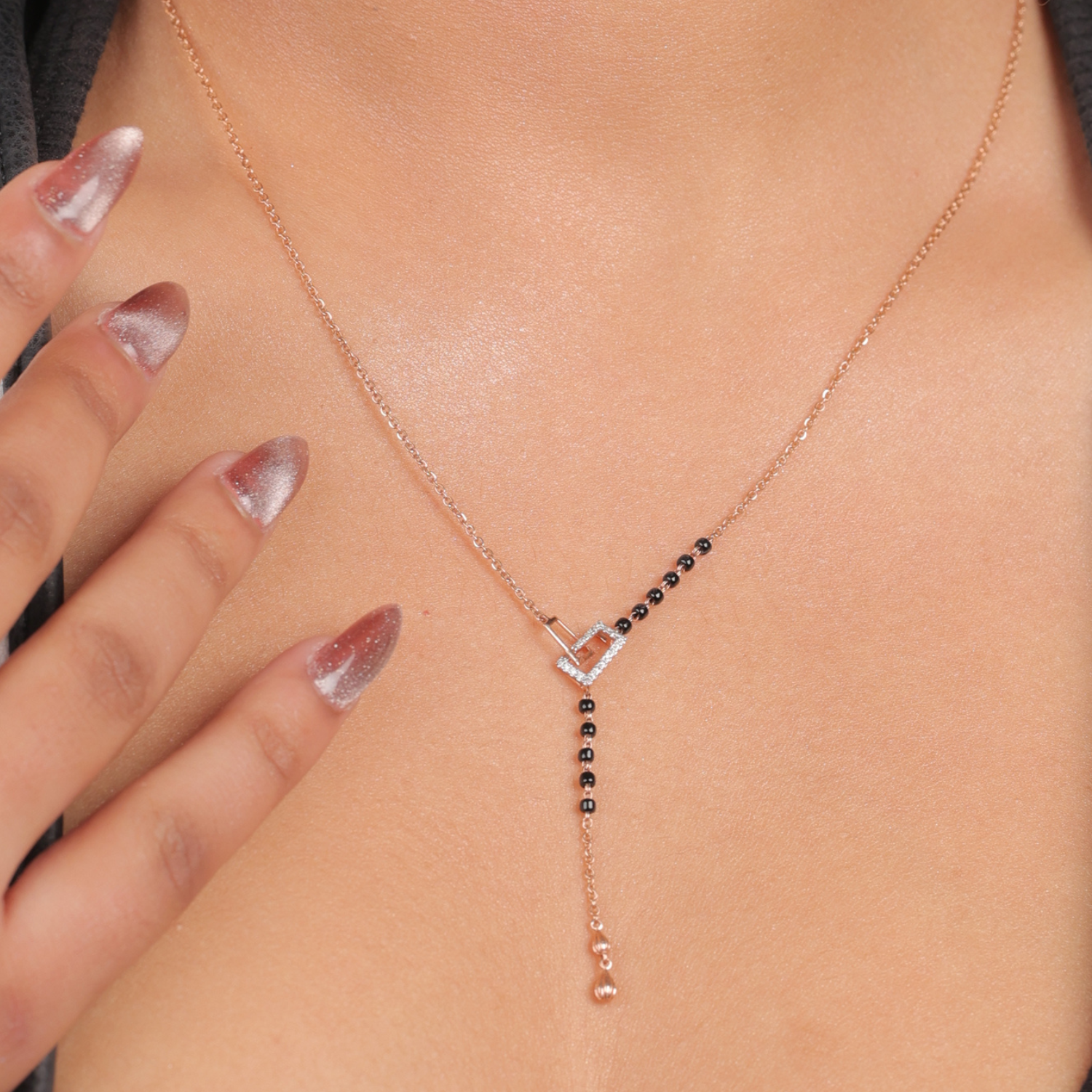 This image showcases the same modern mangalsutra necklace from jnjbymadhuri being worn. The rose gold heart-shaped centerpiece is clearly visible, with a partial segment of traditional black beads leading into a sleek chain and a vertical dangling element. The model's hand, with stylishly manicured nails, gently touches the necklace, emphasizing its elegant and contemporary design.