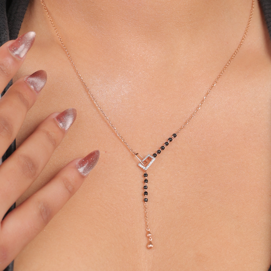 This image showcases the same modern mangalsutra necklace from jnjbymadhuri being worn. The rose gold heart-shaped centerpiece is clearly visible, with a partial segment of traditional black beads leading into a sleek chain and a vertical dangling element. The model's hand, with stylishly manicured nails, gently touches the necklace, emphasizing its elegant and contemporary design.