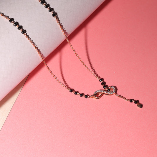 A close-up image of an elegant mangalsutra by jnjbymadhuri, featuring a dainty rose gold chain interlaced with traditional black beads and a stylish infinity motif at the center, placed aesthetically on a curved pastel blue and pink backdrop for a modern and romantic visual appeal.