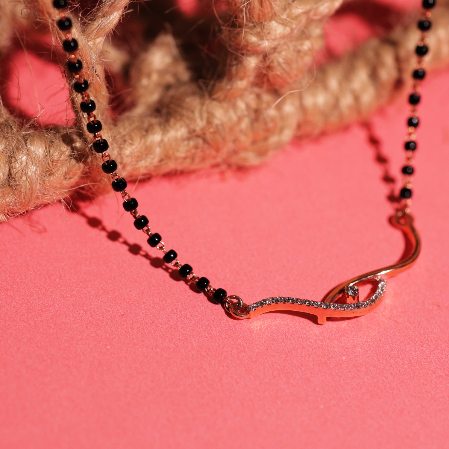 Close-up of a black beaded mangalsutra featuring a twisted gold and diamond pendant, displayed on a coral pink surface with jute twine in the background.