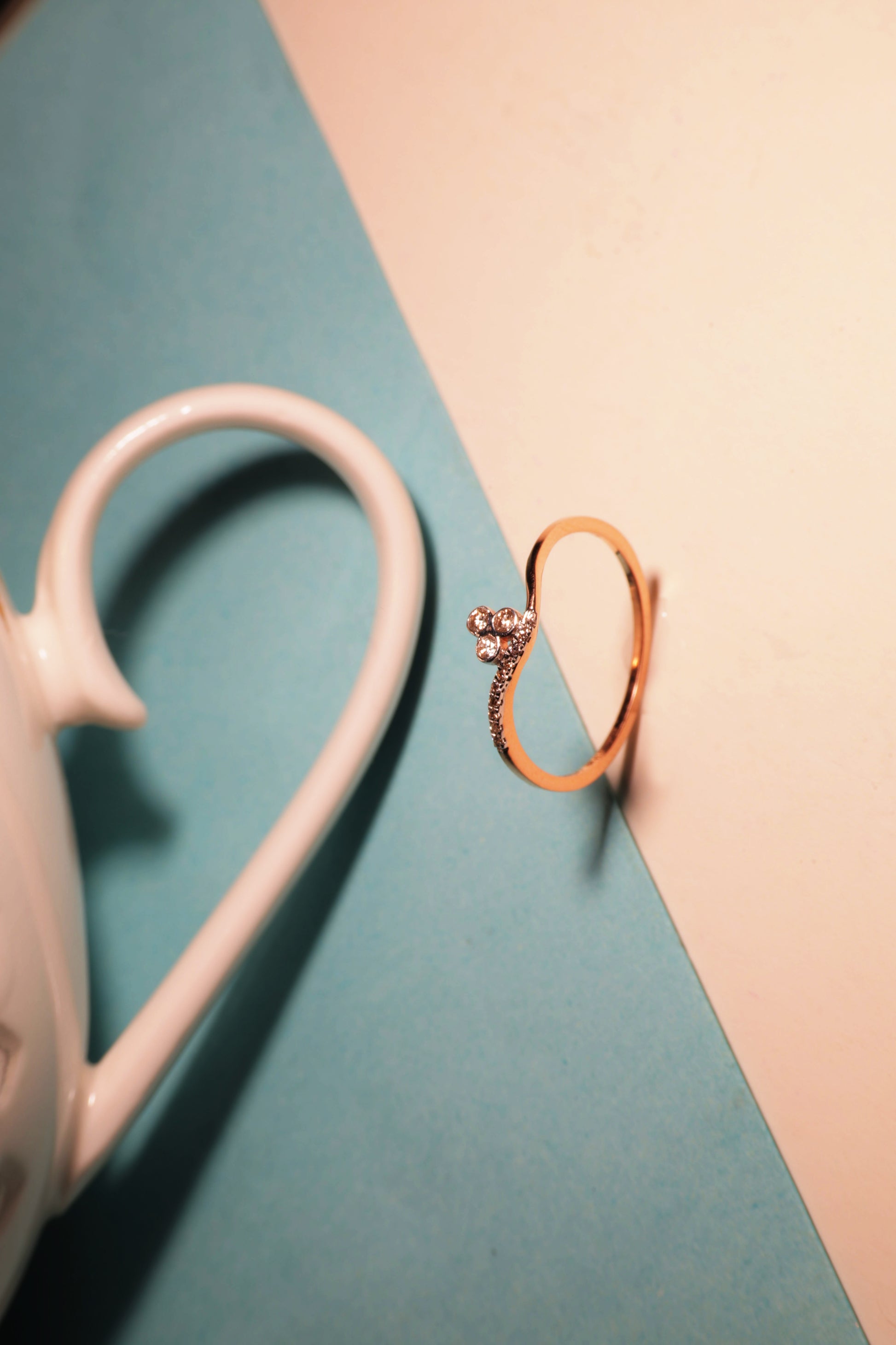A graceful gold ring with a heart-shaped open design and a sparkling diamond accent is artistically balanced on a pastel background beside a ceramic cup, radiating elegance and charm, from jnjbymadhuri.