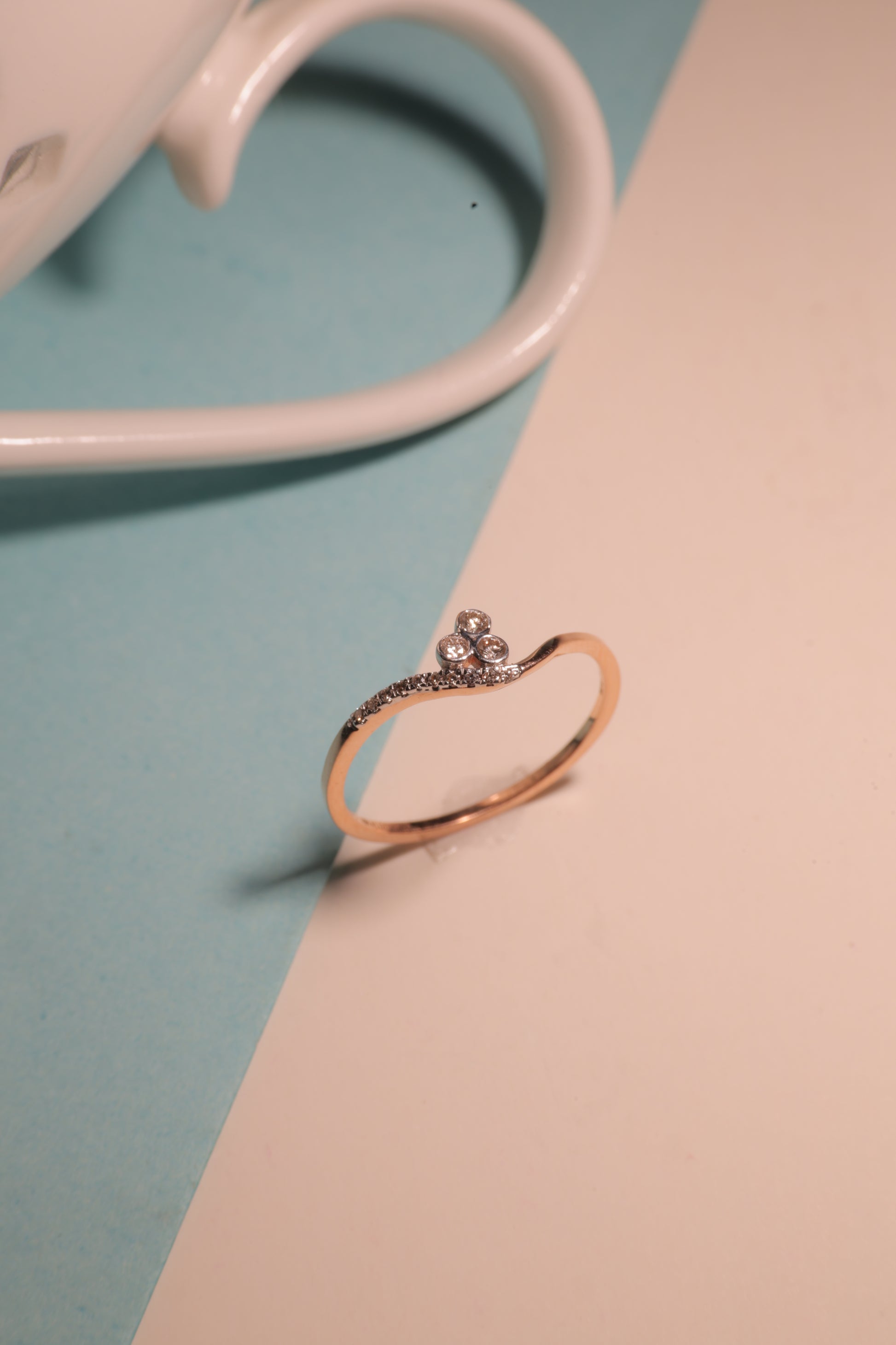 A graceful gold ring with a heart-shaped open design and a sparkling diamond accent is artistically balanced on a pastel background beside a ceramic cup, radiating elegance and charm, from jnjbymadhuri.