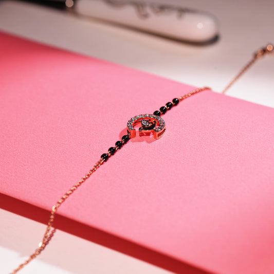 A delicate bracelet by jnjbymadhuri, displayed on a pink box, featuring a rose gold bird motif perched inside a diamond-studded semicircle. The elegant chain is accented with black beads, blending charm and sophistication in a graceful design.