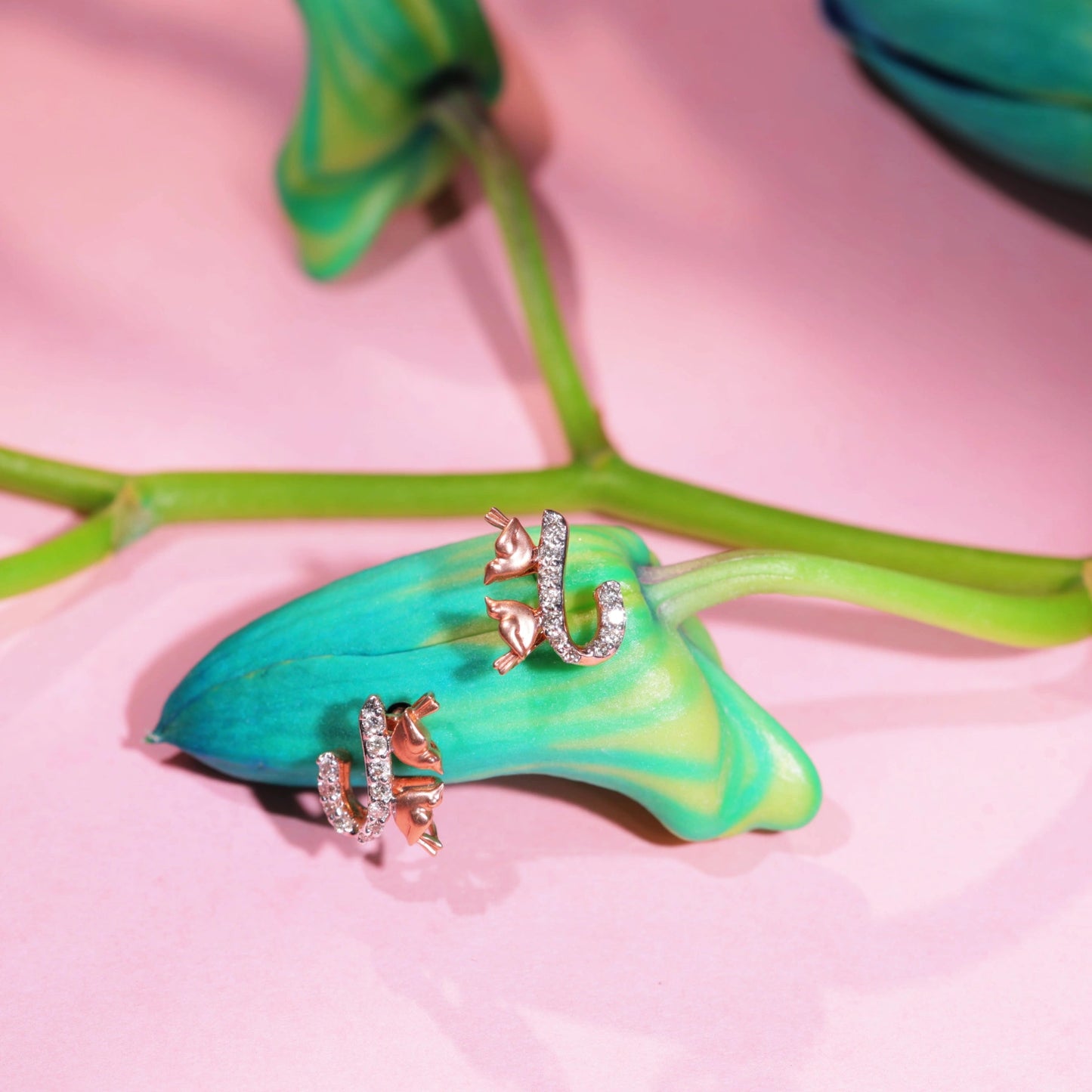 A pair of elegant rose gold earrings from jnjbymadhuri, featuring a delicate cat-and-horseshoe design with shimmering white stones, beautifully styled on a vibrant green bud and stem against a soft pink background, highlighting their whimsical charm and luxurious appeal.