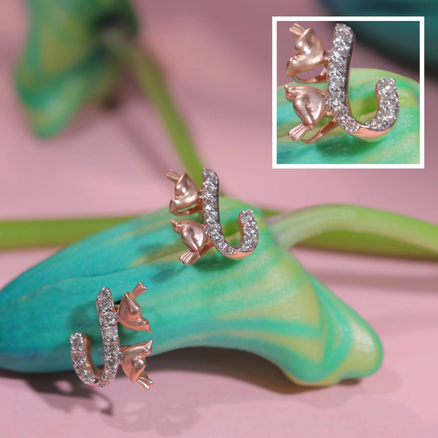 A whimsical pair of rose gold earrings by jnjbymadhuri, designed in the shape of a cat perched on a diamond-studded horseshoe, artfully displayed on a green and teal bud with a soft pink backdrop and a magnified inset showcasing the intricate detailing.