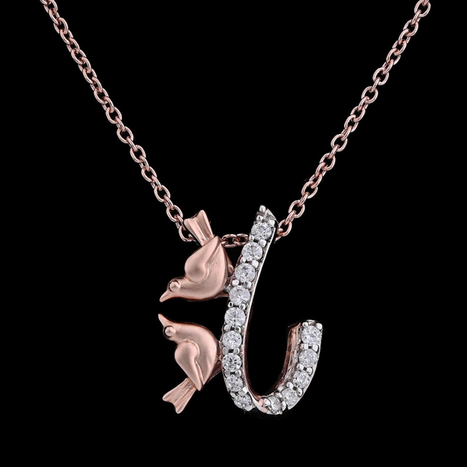 Rose gold necklace featuring a pendant shaped like the letter 'J', embellished with small white stones and two bird motifs perched on the letter, displayed against a black background.