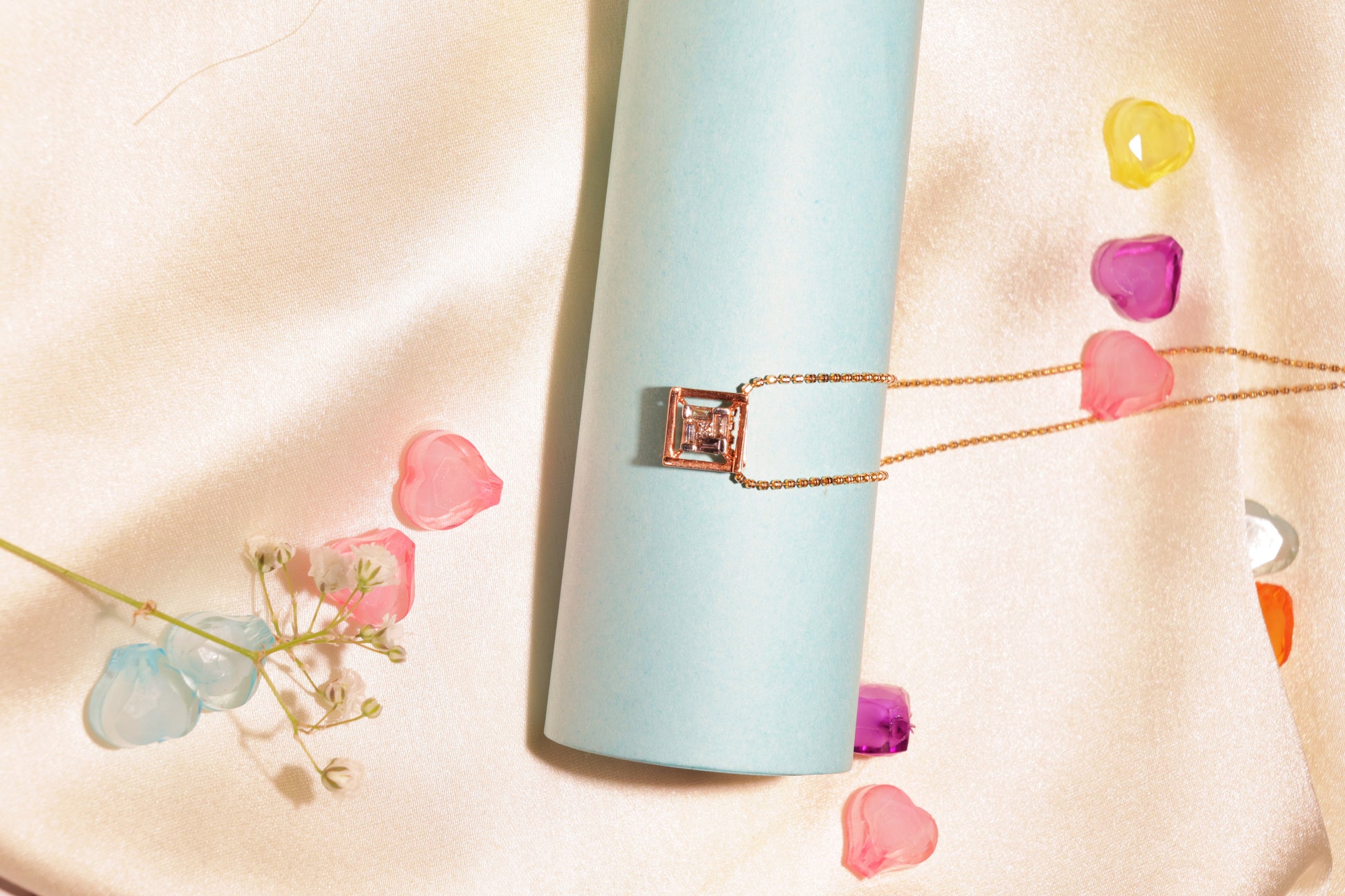 Elegant square diamond pendant necklace by jnjbymadhuri displayed on a light blue cylindrical stand, surrounded by pastel flower petals on a soft satin background.