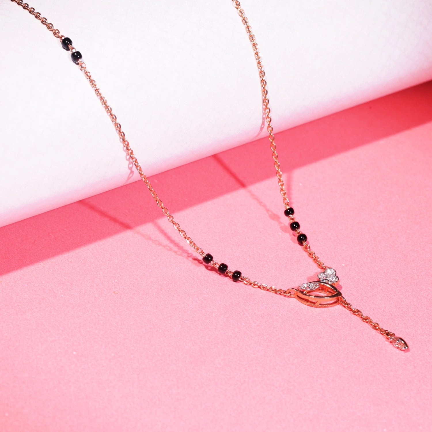 Elegant mangalsutra with a rose gold chain and black bead accents, featuring a central knot-style pendant with a vertical dangling drop, displayed on a vibrant pink background.