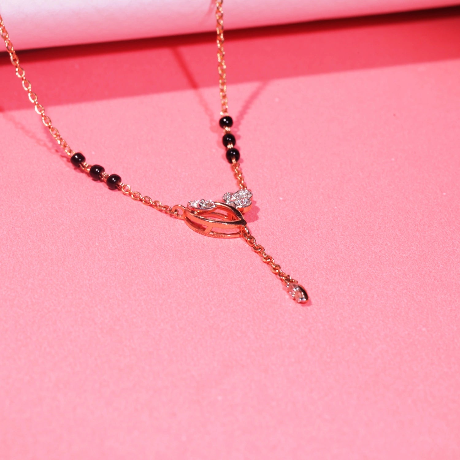 Elegant mangalsutra with a rose gold chain and black bead accents, featuring a central knot-style pendant with a vertical dangling drop, displayed on a vibrant pink background.