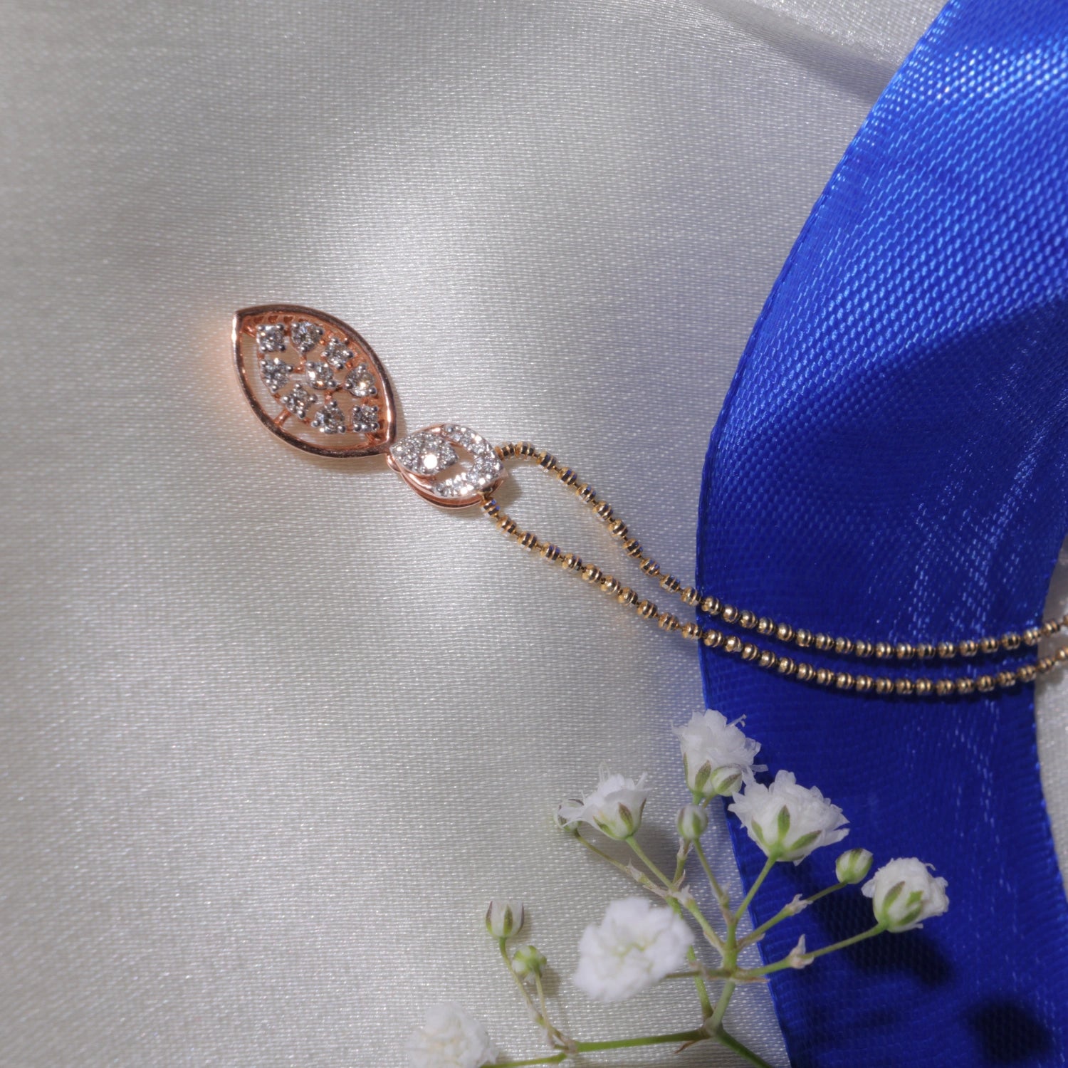 Elegant marquise-shaped rose gold pendant necklace with a diamond cluster, resting on white satin fabric beside a vibrant blue ribbon and delicate white flowers.