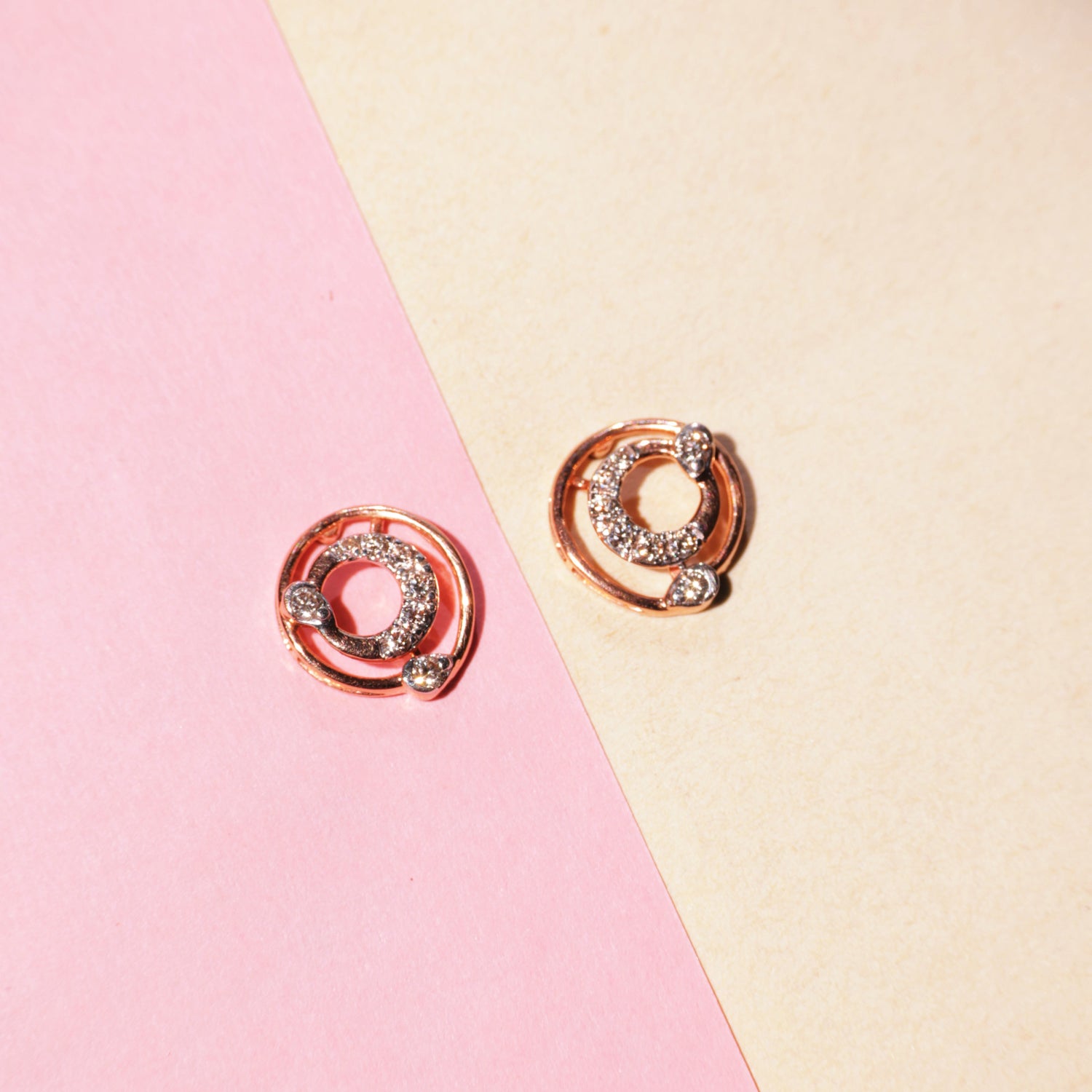 "Rose gold circular earrings with diamond accents arranged on a diagonally split pink and beige background, from jnjbymadhuri."