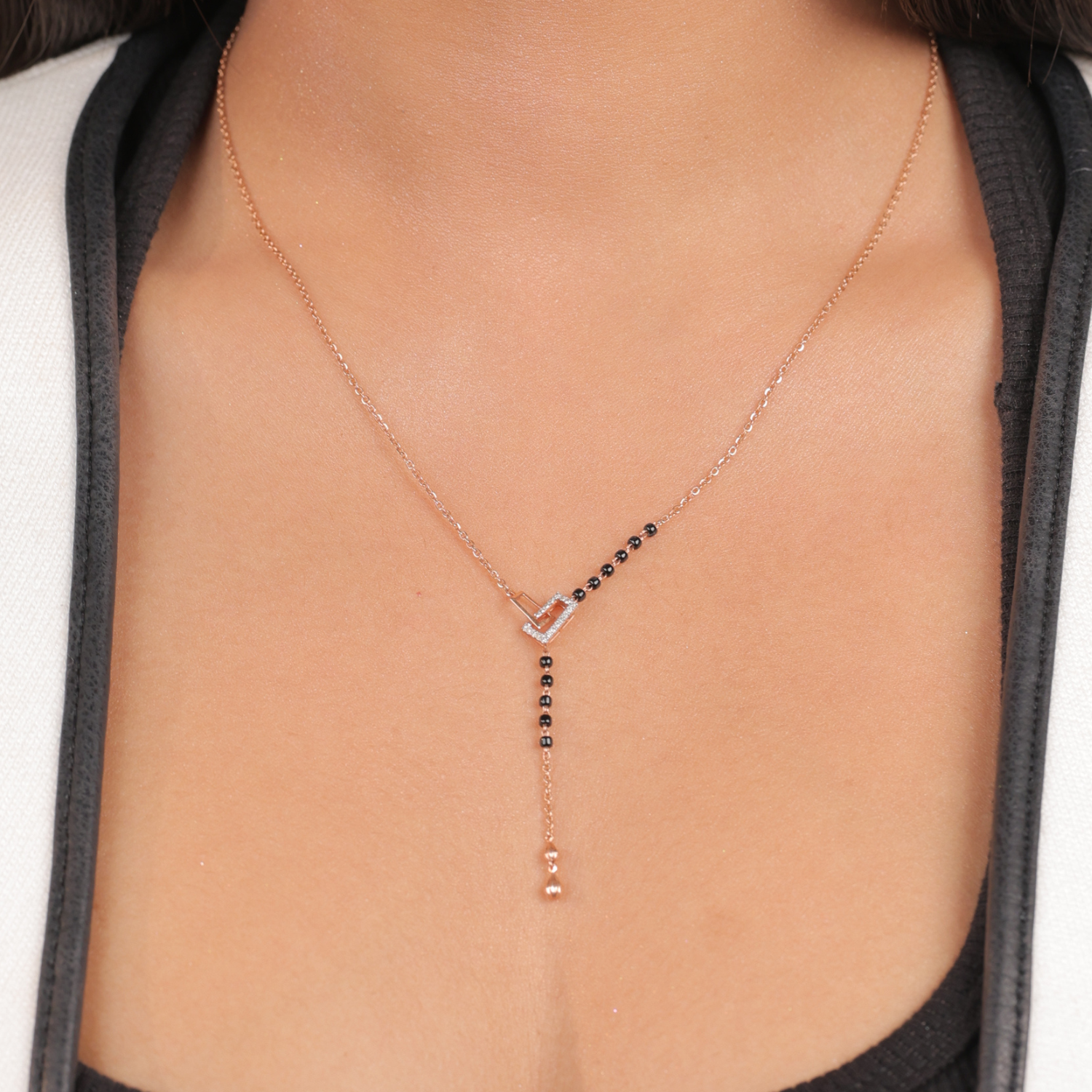 This image showcases the same modern mangalsutra necklace from jnjbymadhuri being worn. The rose gold heart-shaped centerpiece is clearly visible, with a partial segment of traditional black beads leading into a sleek chain and a vertical dangling element. The model's hand, with stylishly manicured nails, gently touches the necklace, emphasizing its elegant and contemporary design.