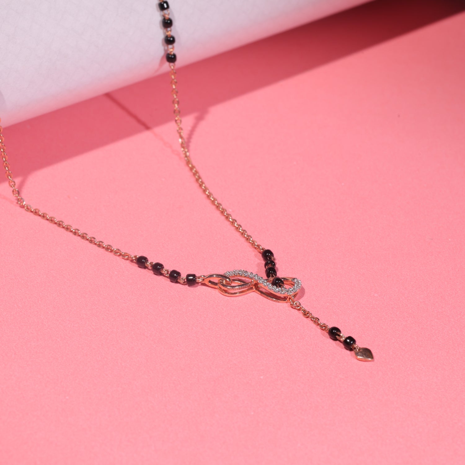 A close-up image of an elegant mangalsutra by jnjbymadhuri, featuring a dainty rose gold chain interlaced with traditional black beads and a stylish infinity motif at the center, placed aesthetically on a curved pastel blue and pink backdrop for a modern and romantic visual appeal.