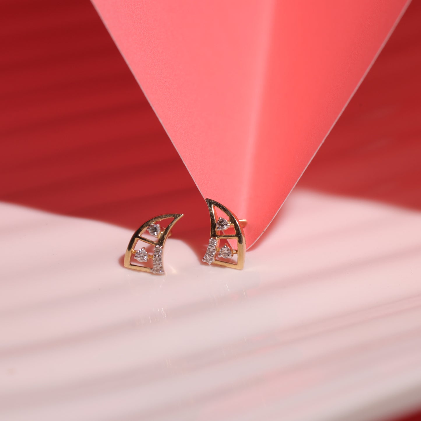 geometric gold and diamond stud earrings by jnjbymadhuri placed under a red origami-style structure, creating a dramatic shadow play on a red and white striped surface.