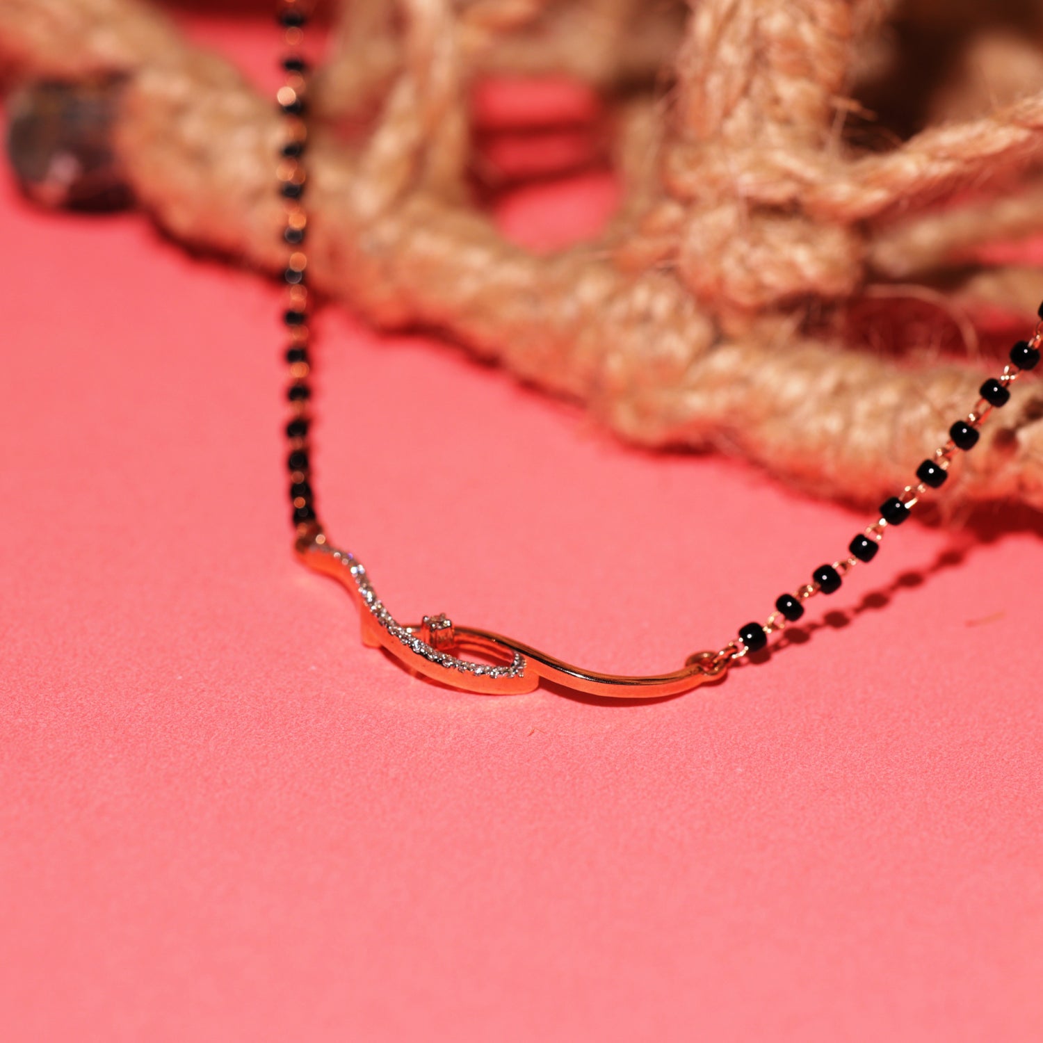 Elegant black beaded mangalsutra with a curved gold and diamond pendant, artistically placed on a coral pink background with natural twine in the backdrop.