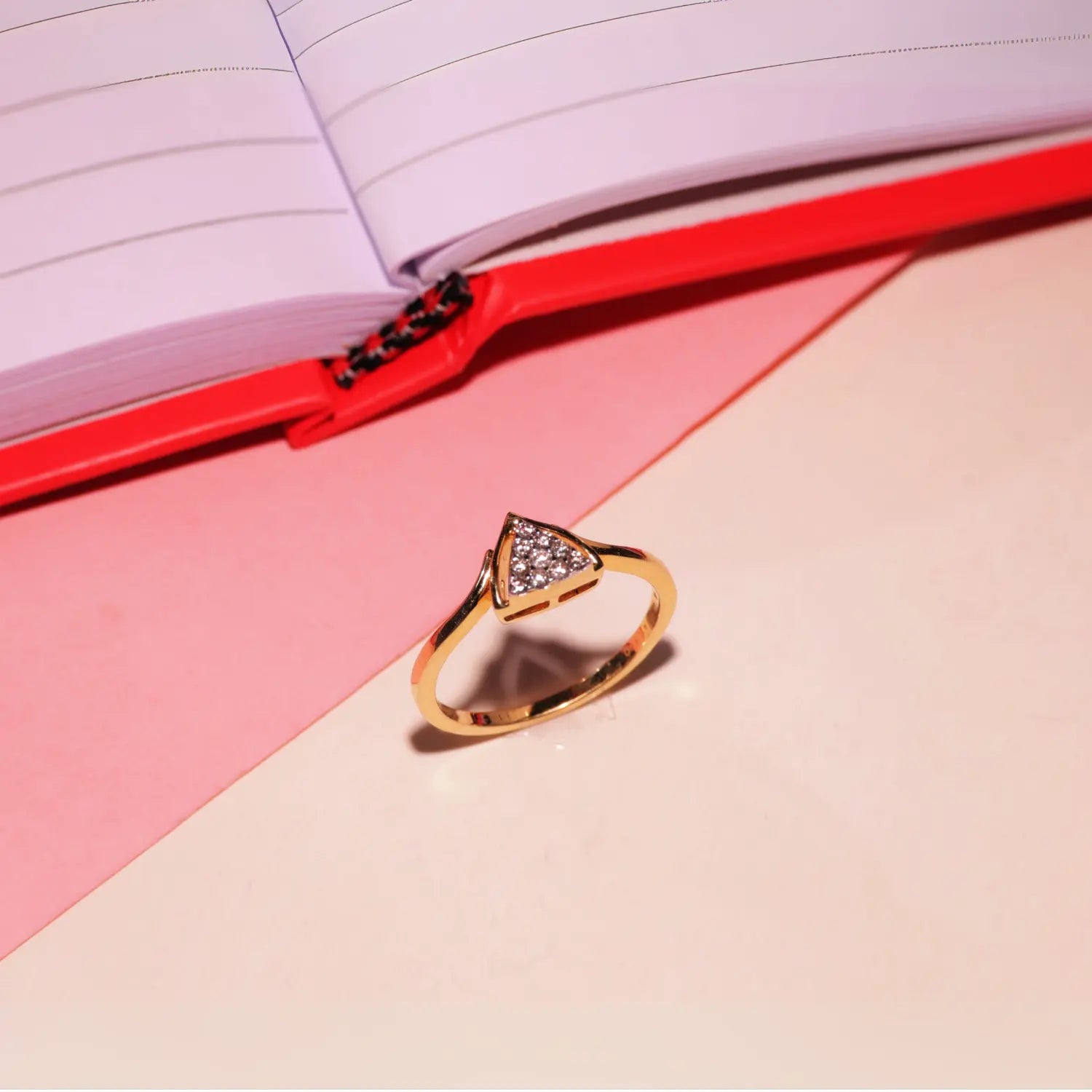A stylish gold ring with a triangular design encrusted with shimmering stones is placed elegantly on a pastel surface near an open notebook, reflecting the modern and chic aesthetic of jnjbymadhuri.