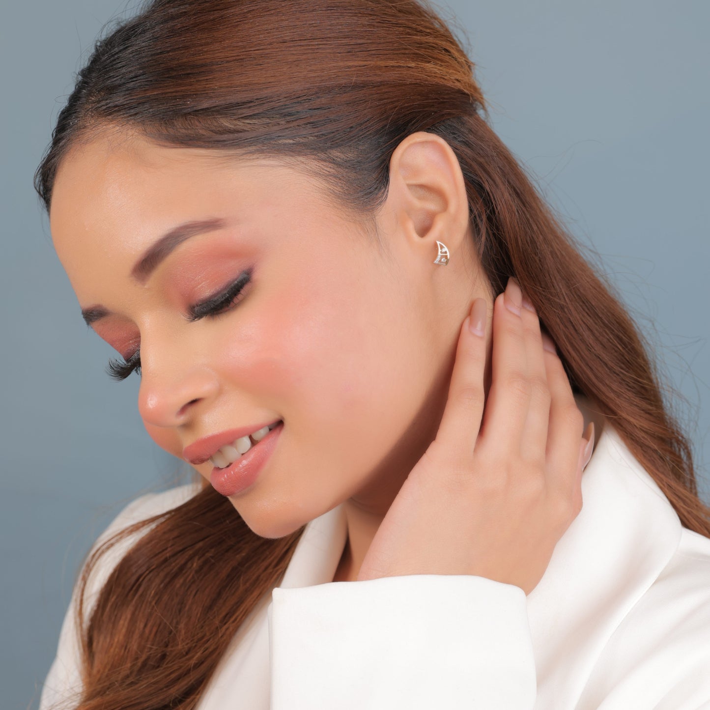 a woman with soft makeup and wavy brown hair wearing minimalist gold and diamond earrings by jnjbymadhuri, smiling gently while touching her neck against a pastel blue background.