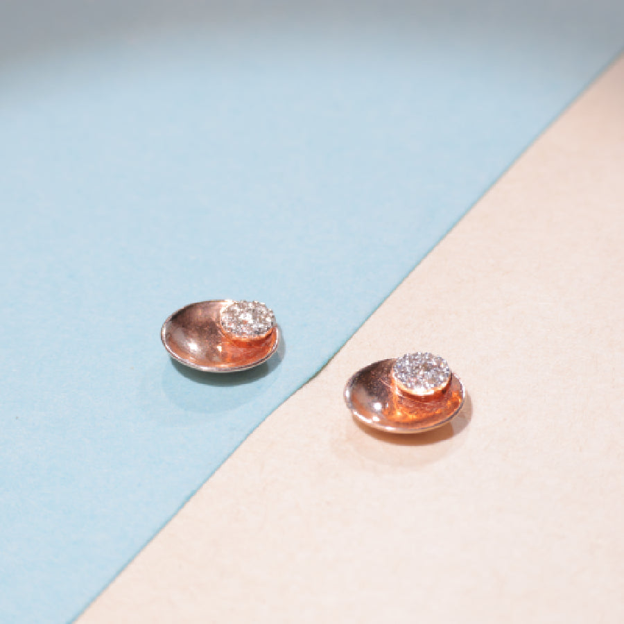 A pair of elegant rose gold stud earrings with diamond-studded tops placed on a pastel blue and cream background. Timeless luxury captured by jnjbymadhuri.