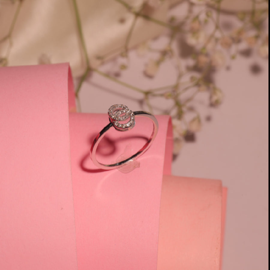 A delicate silver ring with a round, flower-inspired diamond cluster centerpiece is elegantly displayed on soft pink rolled paper, exuding feminine charm and timeless elegance by jnjbymadhuri.
