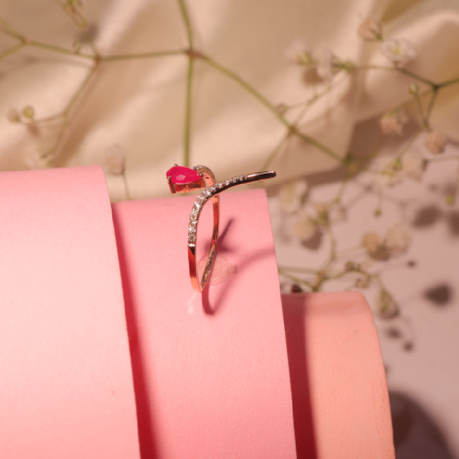 Heart-shaped ruby ring with a diamond-studded curved band set in rose gold, styled elegantly on pink spirals. A timeless piece by jnjbymadhuri.