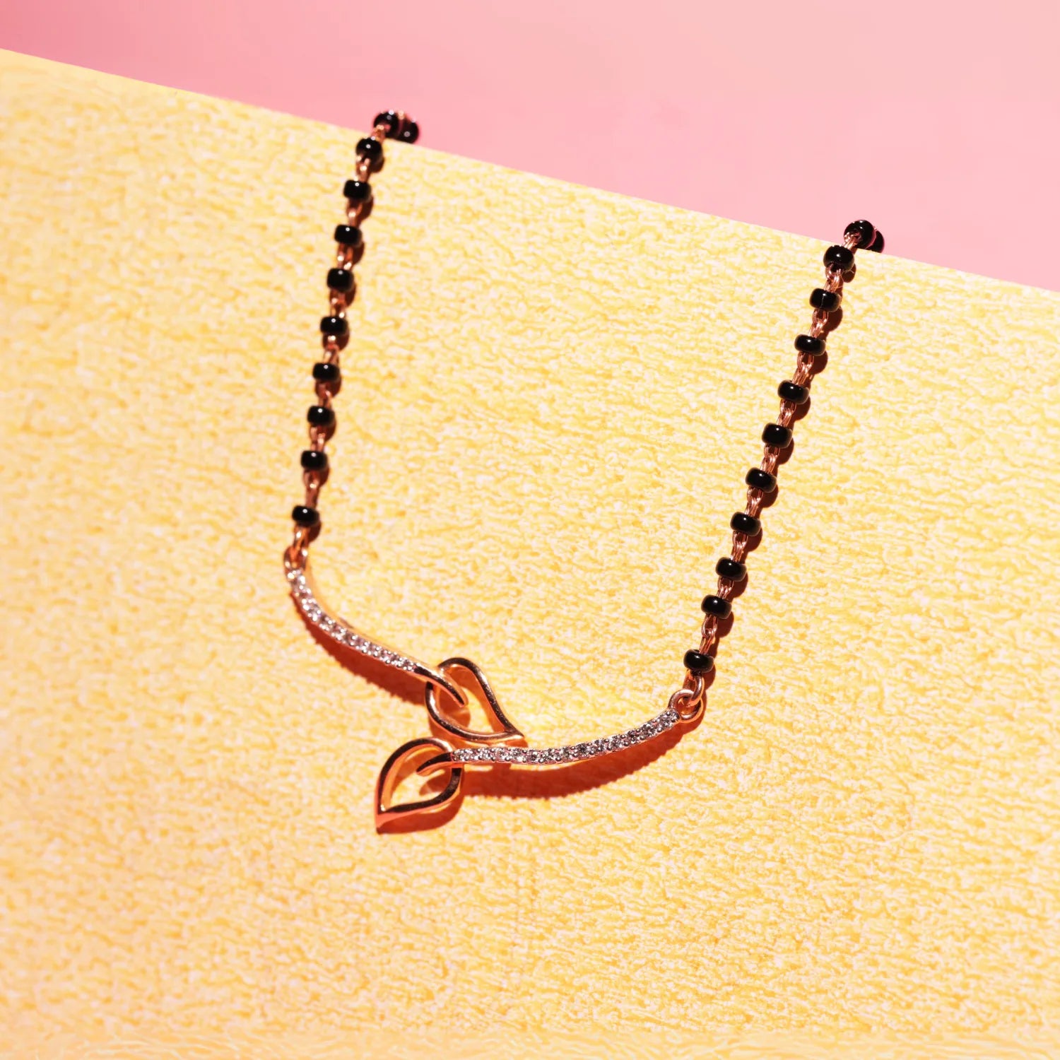 Artistic display of a jnjbymadhuri mangalsutra featuring a twin-leaf gold pendant with a sleek curved design, attached to a traditional black beaded chain, elegantly placed on a pastel pink and beige backdrop for a modern aesthetic.