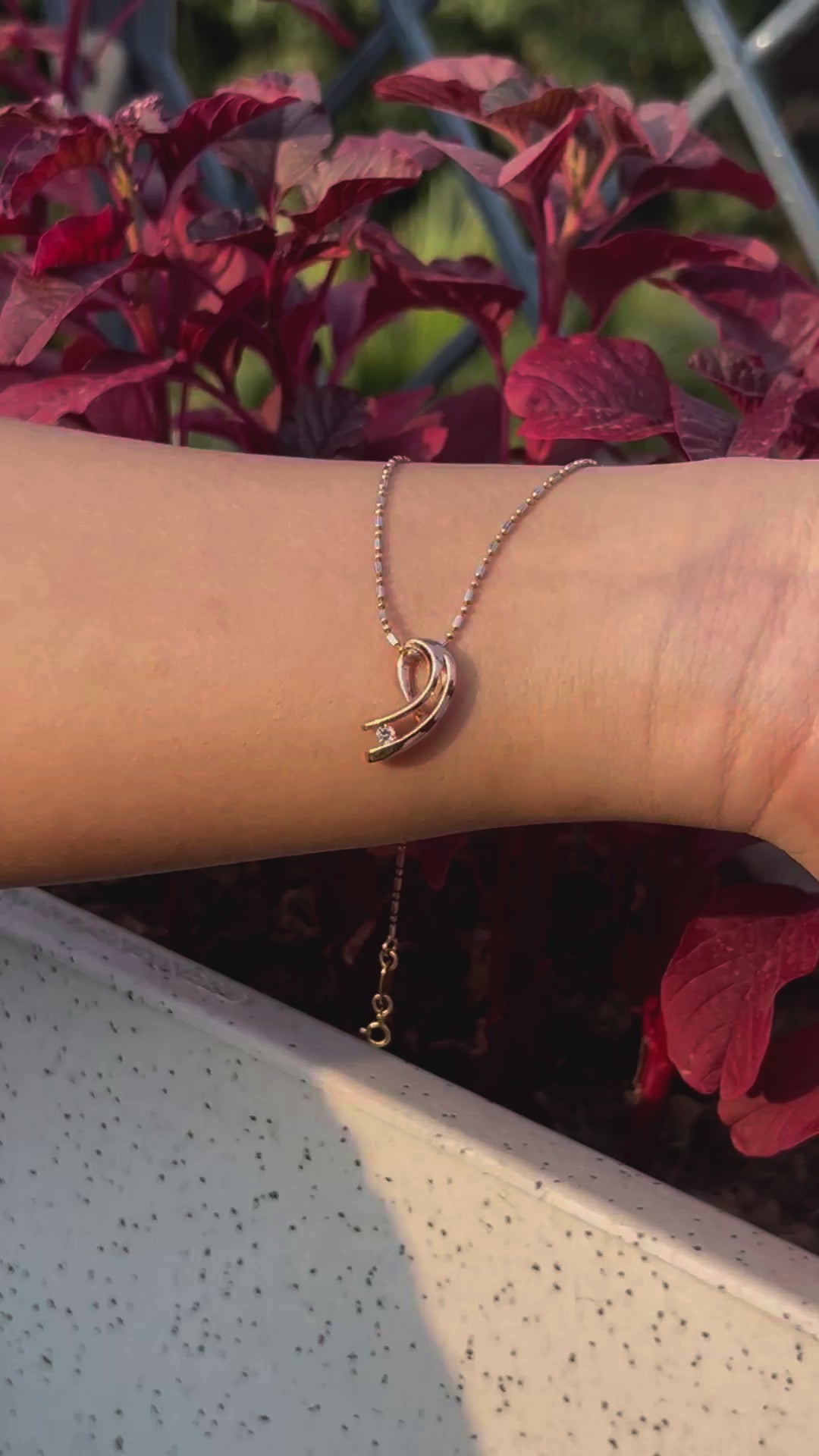 Wrist adorned with a jnjbymadhuri rose gold bracelet featuring a curved pendant and single white stone, showcased against vibrant red foliage in natural daylight