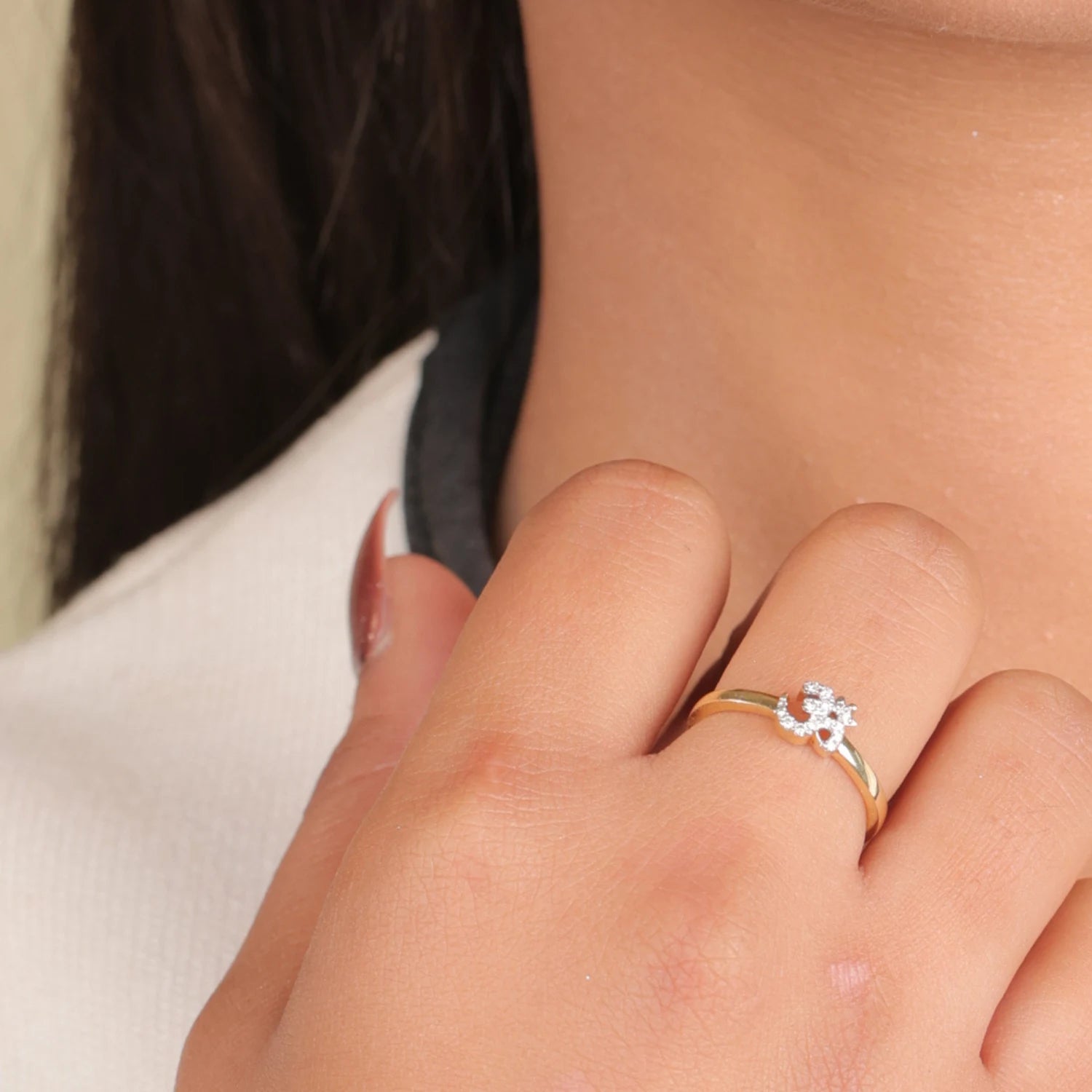 A delicate gold ring featuring a sparkling diamond-studded "Om" symbol is gracefully worn on a woman's finger, complementing her poised hand placement near the neckline, highlighting the ring’s spiritual charm and refined style, from jnjbymadhuri.
