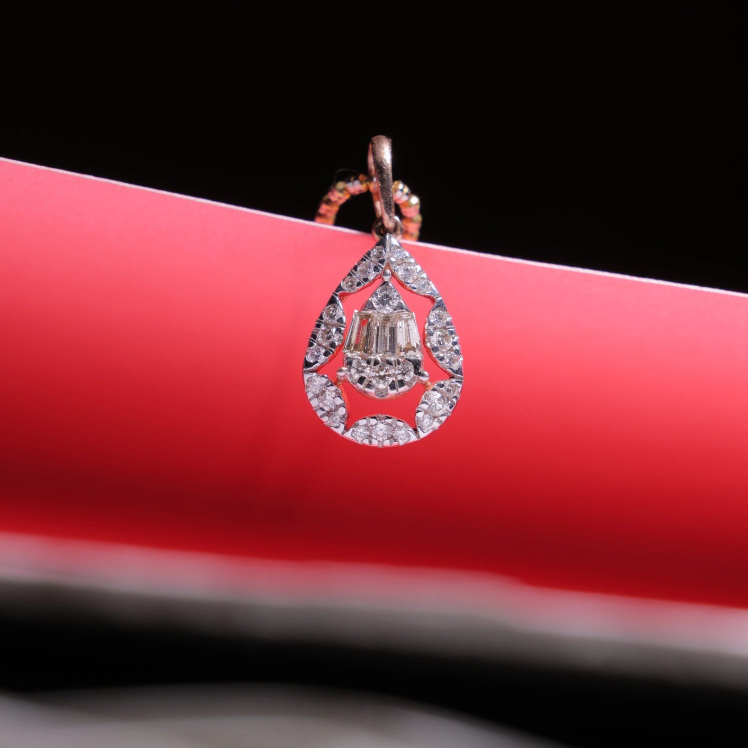 Rose gold teardrop-shaped pendant with sparkling diamonds by jnjbymadhuri, displayed elegantly against a vibrant red and black backdrop.
