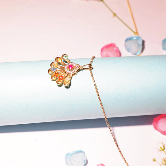 Artistic lifestyle shot of a vibrant Navratna pendant by jnjbymadhuri, displayed on a delicate gold chain over a pastel blue cylinder, surrounded by soft-focus rose petals and a dreamy pink backdrop—highlighting the pendant’s nine colorful gemstones and elegant fan-shaped gold frame.
