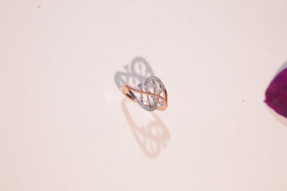 Rose gold ring with a swirling, open design and embedded diamonds, casting an elegant shadow on a pale surface with a purple object in the corner.
