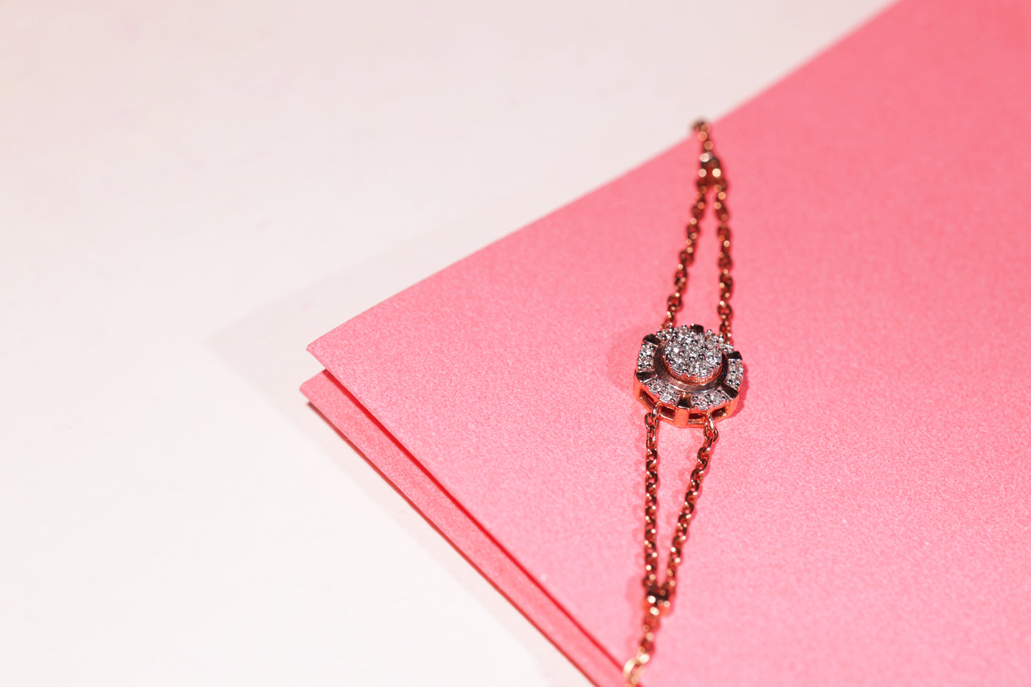 A stunning dual-chain rose gold bracelet by jnjbymadhuri, featuring a central floral motif encrusted with sparkling stones, elegantly displayed against a pink textured background—an embodiment of feminine grace and refined craftsmanship.
