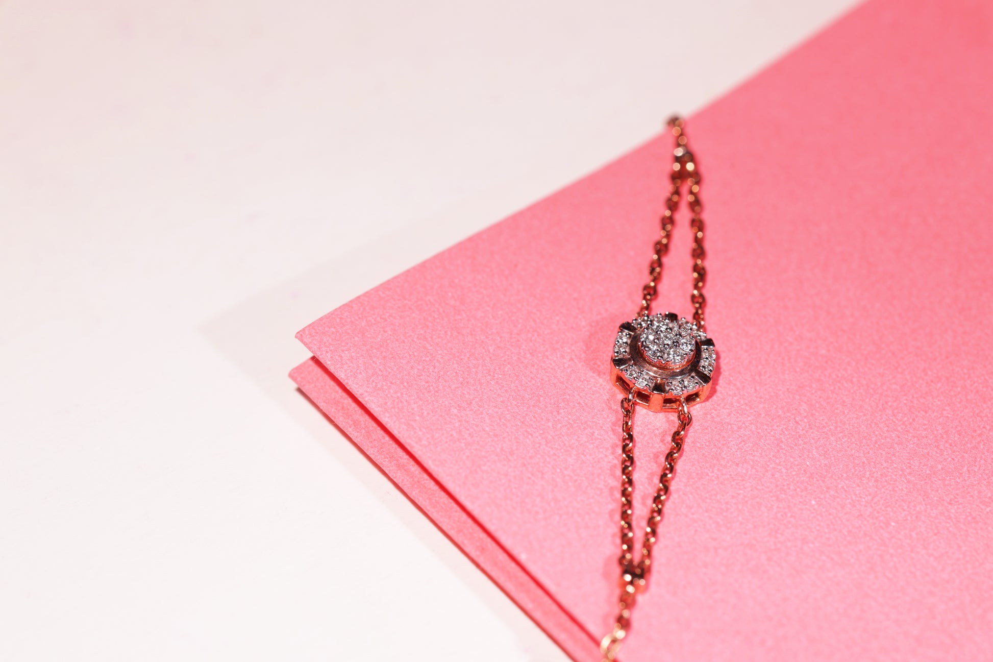 A stunning dual-chain rose gold bracelet by jnjbymadhuri, featuring a central floral motif encrusted with sparkling stones, elegantly displayed against a pink textured background—an embodiment of feminine grace and refined craftsmanship.
