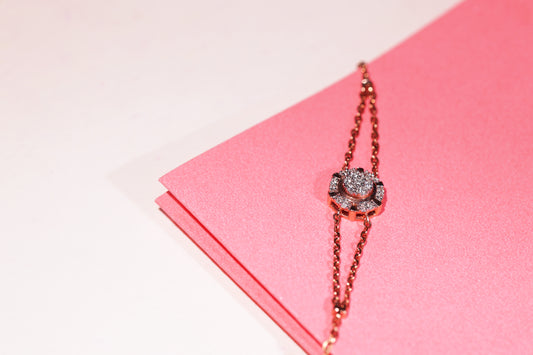 A stunning dual-chain rose gold bracelet by jnjbymadhuri, featuring a central floral motif encrusted with sparkling stones, elegantly displayed against a pink textured background—an embodiment of feminine grace and refined craftsmanship.
