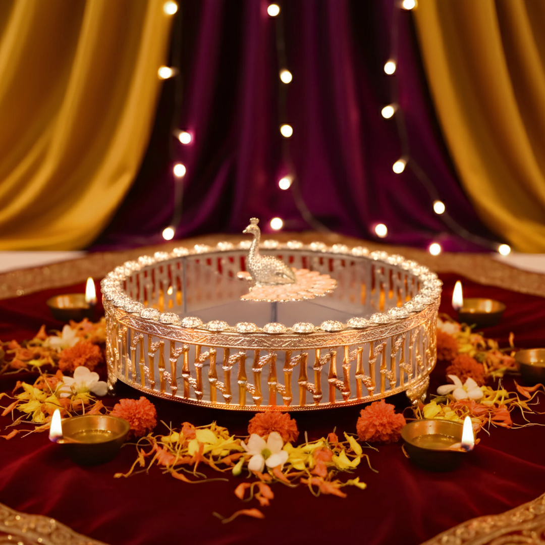 Decorative lamp with peacock design on a table with flowers and candles, against a draped curtain background.