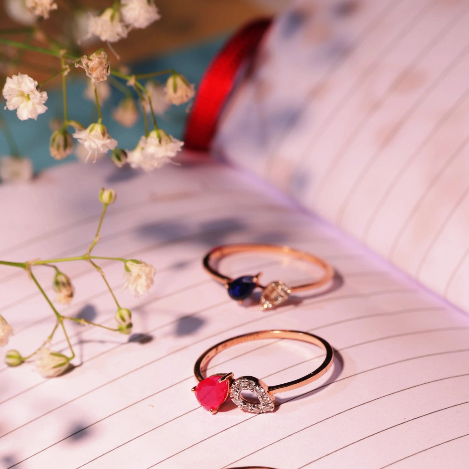 Three elegant gemstone rings by jnjbymadhuri, featuring red, blue, and clear stones, placed aesthetically on an open notebook with delicate white flowers.
