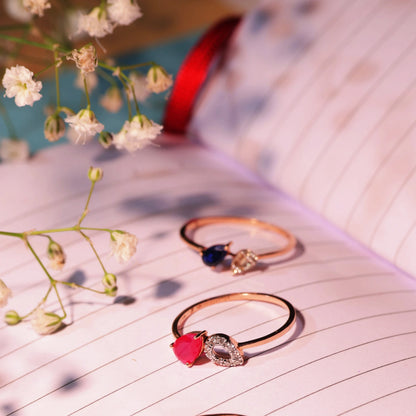 Three elegant gemstone rings by jnjbymadhuri, featuring red, blue, and clear stones, placed aesthetically on an open notebook with delicate white flowers.

