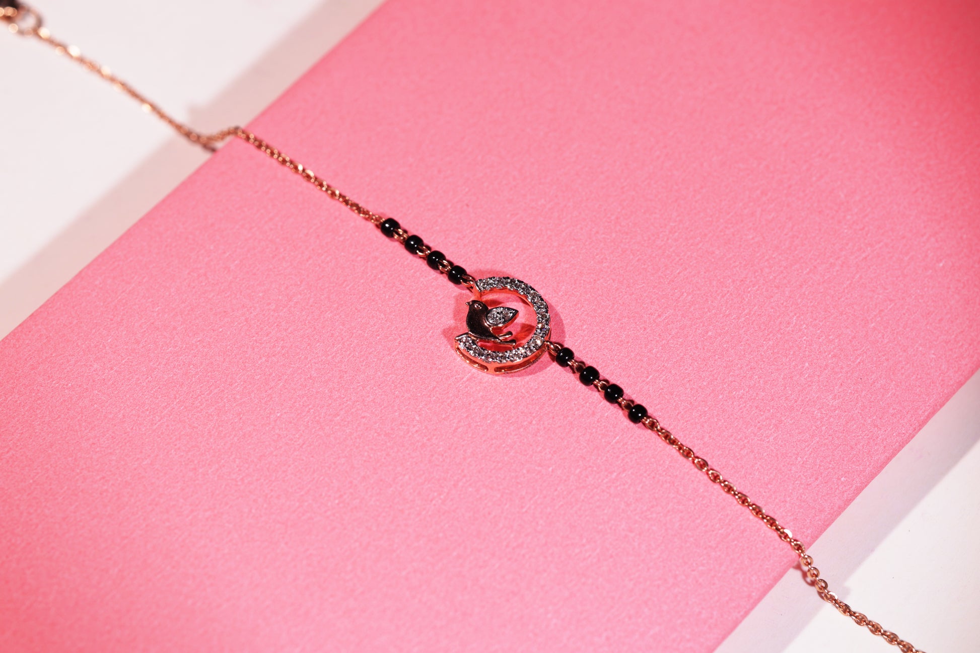 A delicate bracelet by jnjbymadhuri, displayed on a pink box, featuring a rose gold bird motif perched inside a diamond-studded semicircle. The elegant chain is accented with black beads, blending charm and sophistication in a graceful design.
