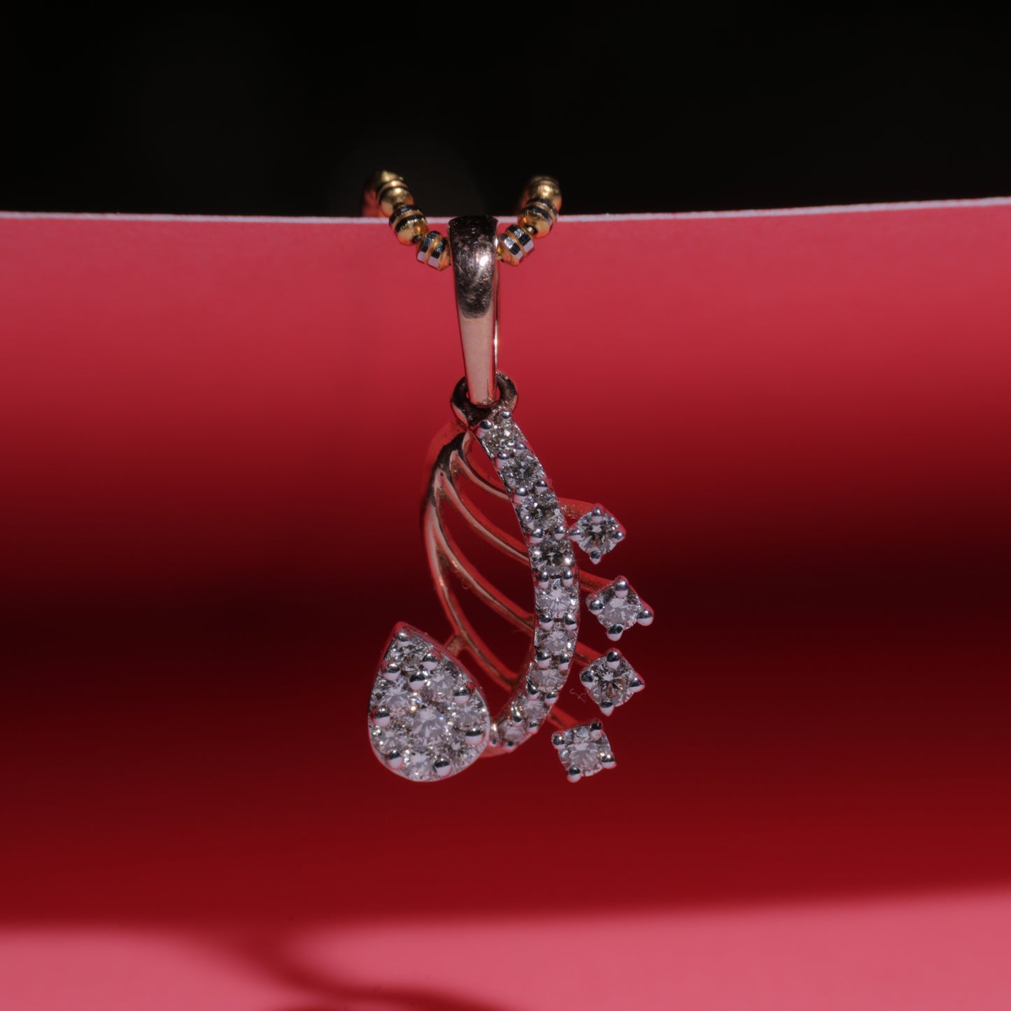 Rose gold paisley-shaped pendant by jnjbymadhuri, featuring intricate design with embedded white stones, displayed on a vibrant red background with golden chain loops.
