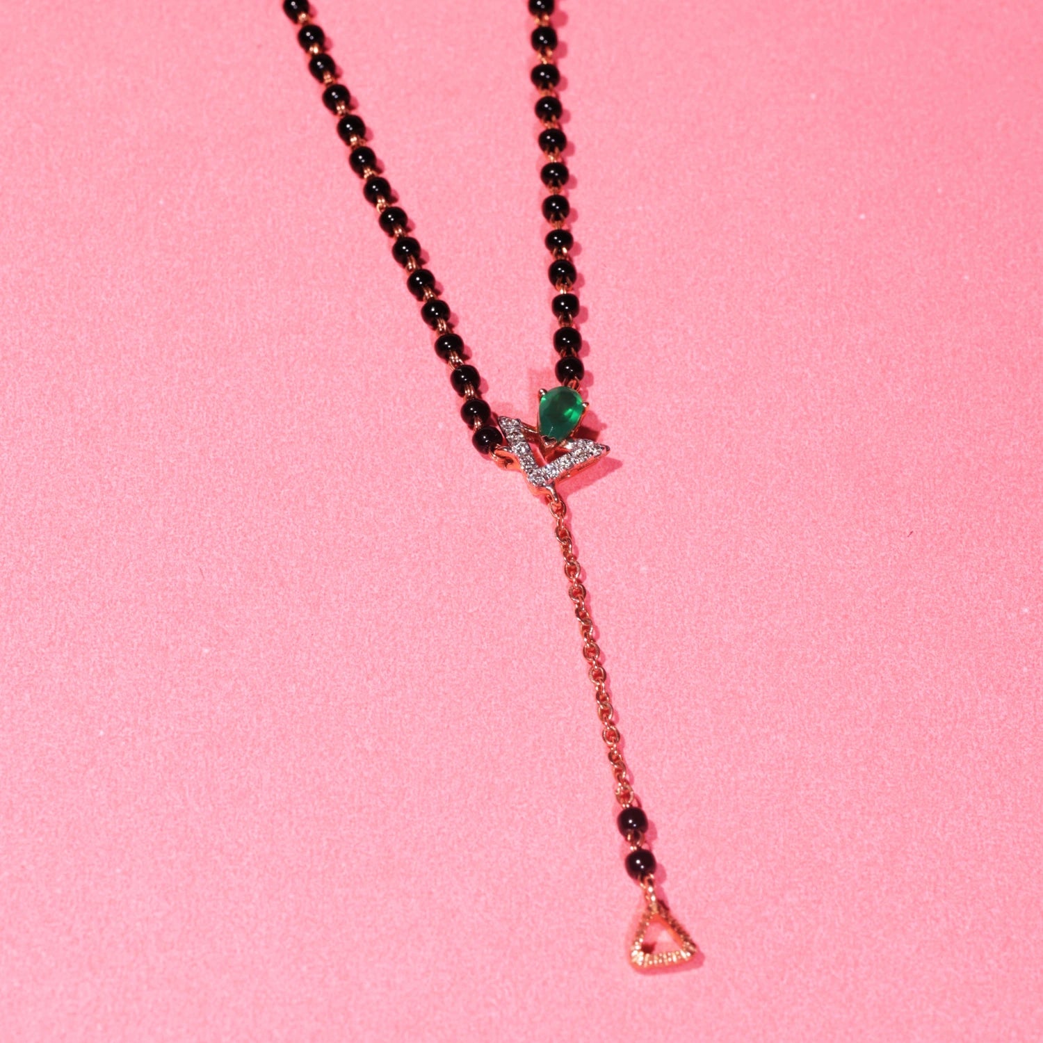 Stylish jnjbymadhuri mangalsutra displayed against a pink background, showcasing a modern black bead chain, an eye-catching emerald green stone, and a distinctive gold drop pendant with delicate detailing for a contemporary yet traditional look.
