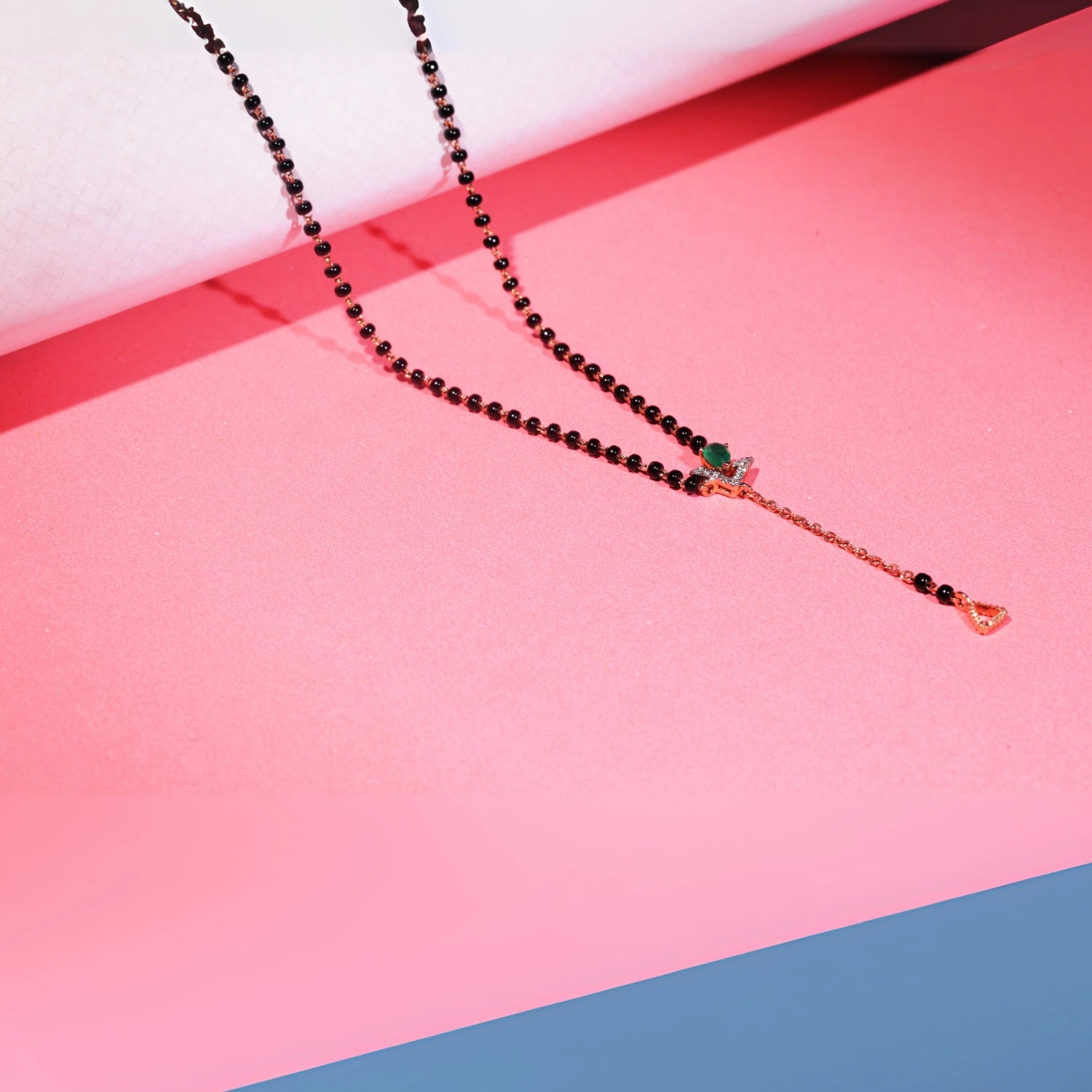 Stylish jnjbymadhuri mangalsutra displayed against a pink background, showcasing a modern black bead chain, an eye-catching emerald green stone, and a distinctive gold drop pendant with delicate detailing for a contemporary yet traditional look.

