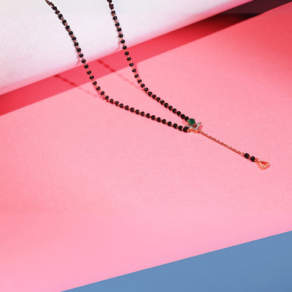 Stylish jnjbymadhuri mangalsutra displayed against a pink background, showcasing a modern black bead chain, an eye-catching emerald green stone, and a distinctive gold drop pendant with delicate detailing for a contemporary yet traditional look.
