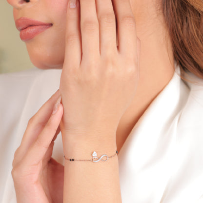 Delicate rose gold infinity bracelet with a dangling heart charm, worn elegantly on the wrist for a minimal yet romantic look. A refined everyday piece by jnjbymadhuri.
