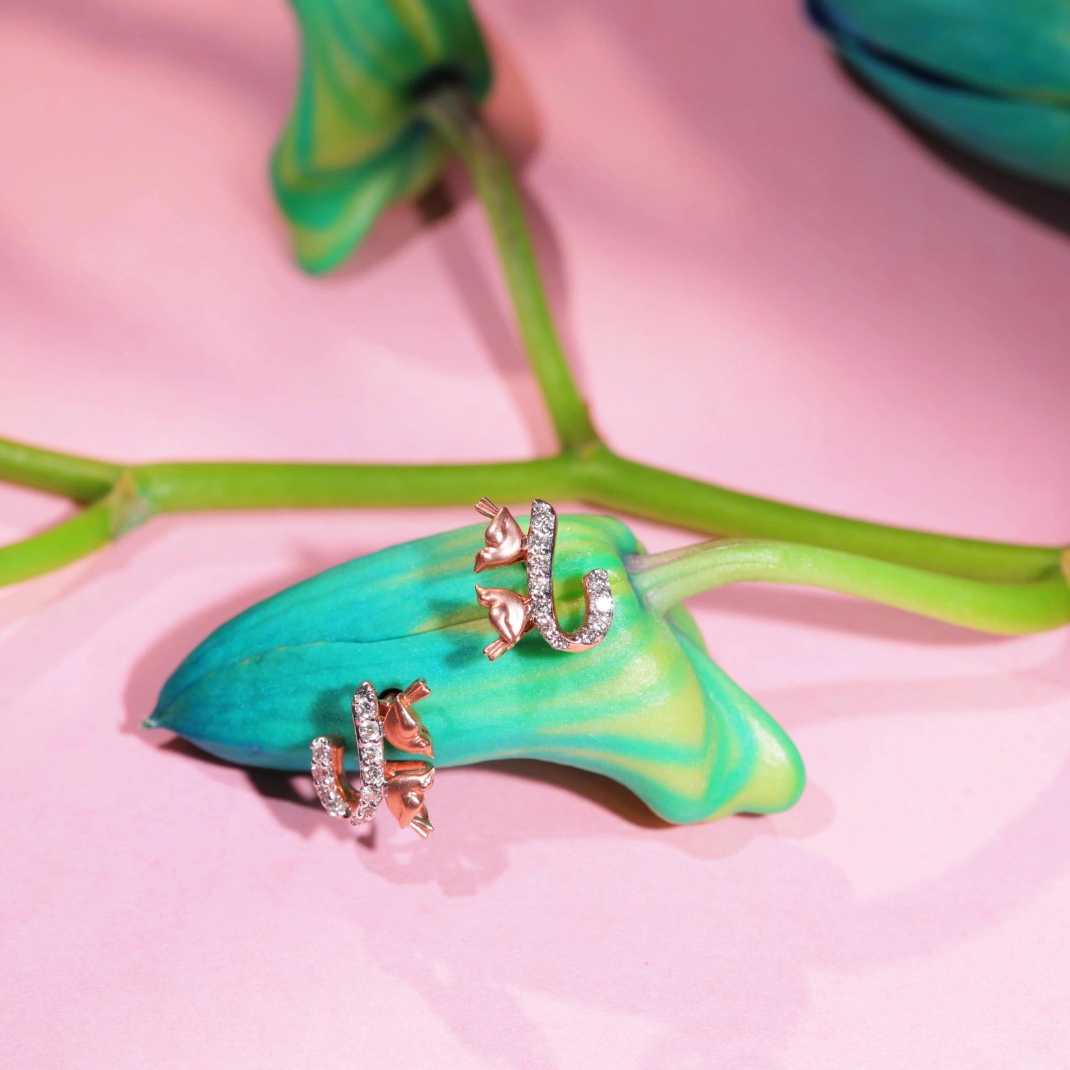 A pair of elegant rose gold earrings from jnjbymadhuri, featuring a delicate cat-and-horseshoe design with shimmering white stones, beautifully styled on a vibrant green bud and stem against a soft pink background, highlighting their whimsical charm and luxurious appeal.
