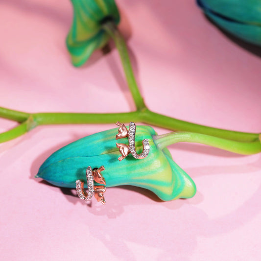 A pair of elegant rose gold earrings from jnjbymadhuri, featuring a delicate cat-and-horseshoe design with shimmering white stones, beautifully styled on a vibrant green bud and stem against a soft pink background, highlighting their whimsical charm and luxurious appeal.
