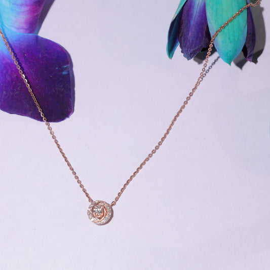 Elegant rose gold necklace with a spiral diamond pendant by jnjbymadhuri, artistically displayed against a pastel background with vibrant purple and teal petals.
