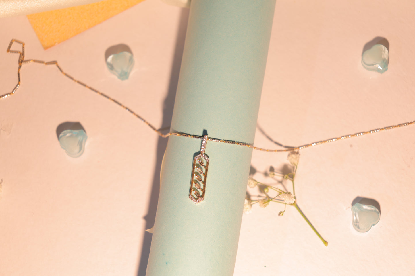 Gold pendant necklace with a vertical hexagonal frame and chevron diamond pattern displayed on a pastel setup with blue cylindrical props and decorative baby’s breath flowers.
