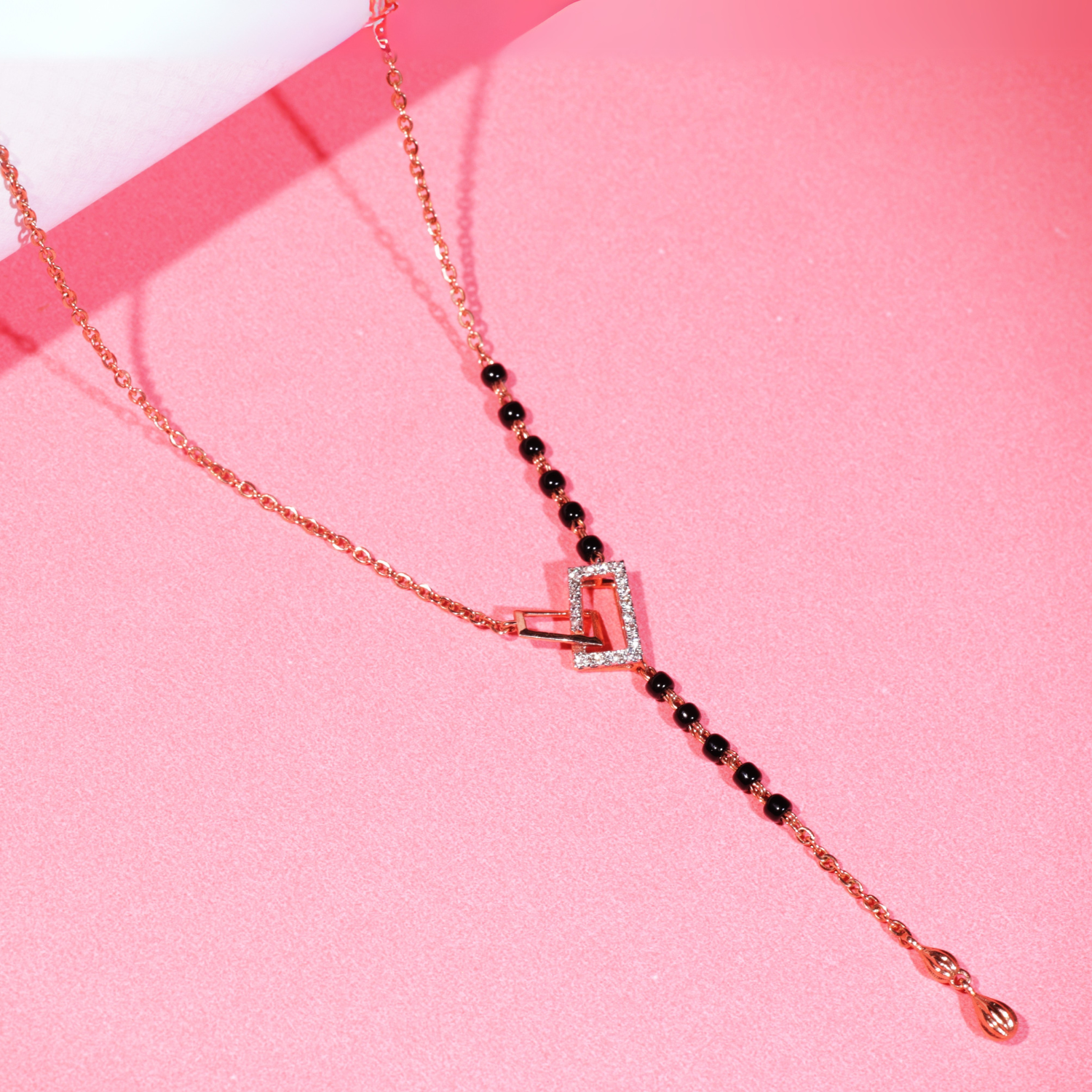 This image presents a close-up, stylized flat-lay of the same modern mangalsutra with a heart-shaped pendant. Set against a pink background, the necklace’s details stand out clearly—rose gold chain, a partial segment of traditional black beads, and a Y-drop design ending with a small dangling charm. The heart centerpiece is adorned with subtle embellishments, giving the piece a blend of tradition and contemporary elegance.
