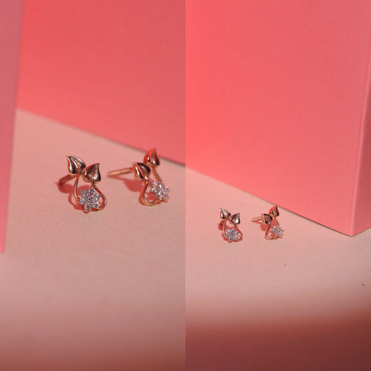 Charming bow-shaped earrings by jnjbymaduri, adorned with sparkling stones and a heart-shaped drop, elegantly displayed against a soft pink backdrop for a playful yet sophisticated look.
